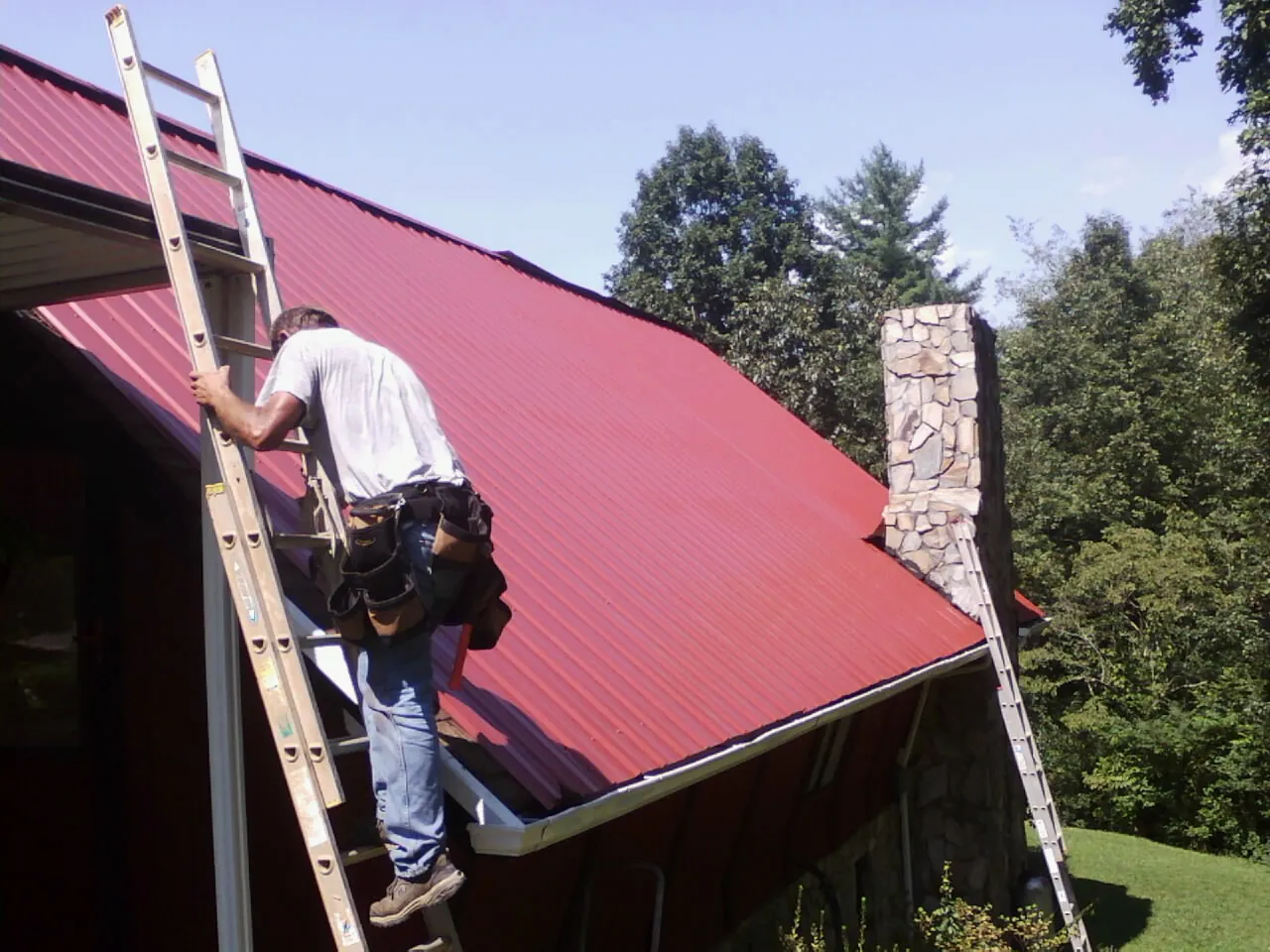 Roofer on a ladder beside a red metal roof and stone chimney — Soffit & Fascia Repair by Roofing Brothers in Crisfield